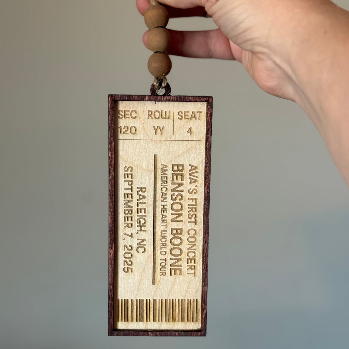 A hand holds a wooden concert ticket ornament featuring engraved text for someone's first concert, with a beaded hanging cord against a plain gray background.
