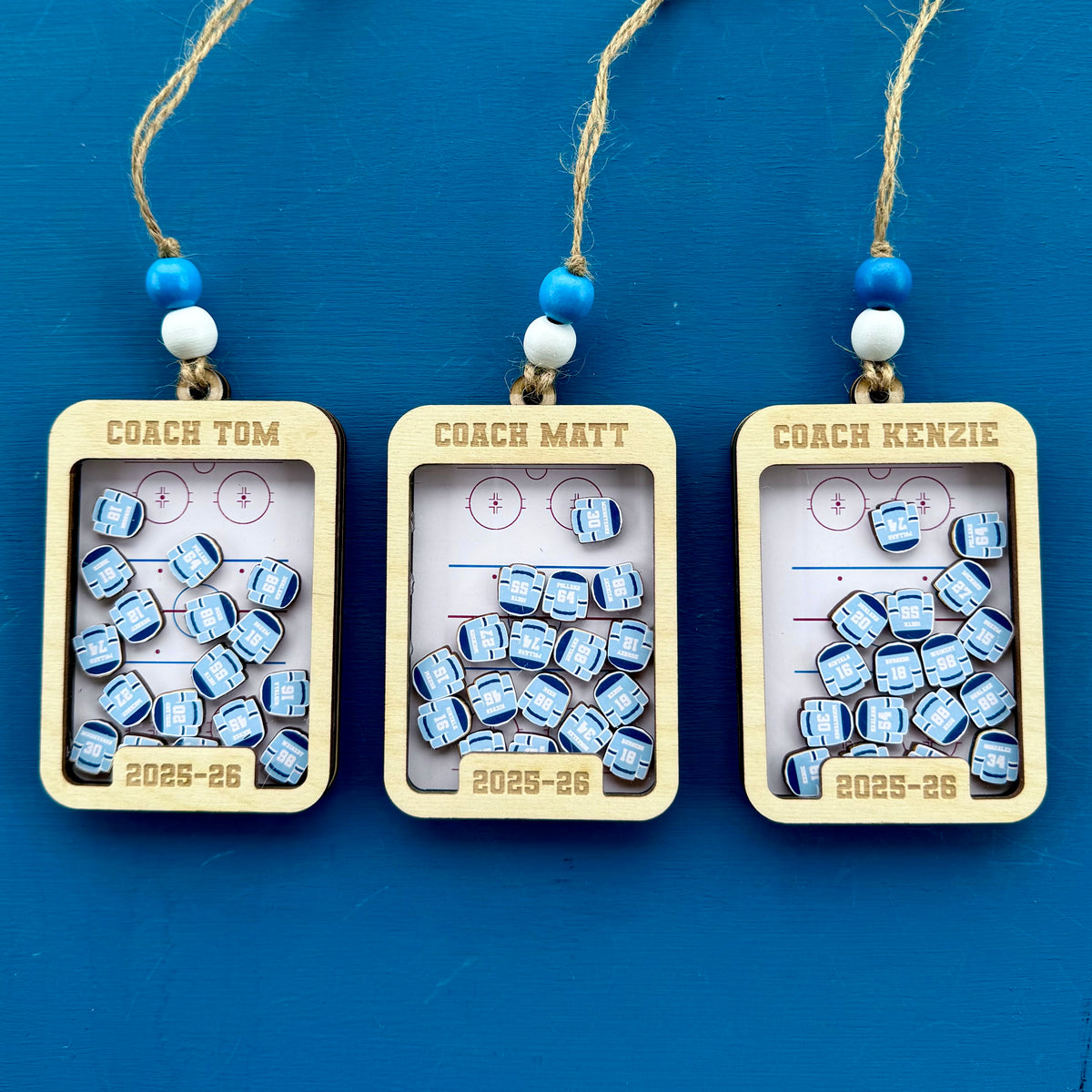 Three personalized wooden ornaments are arranged side by side on a blue background. Each ornament has a transparent window showing small blue-and-white wooden pieces with an image of a hockey jersey with play name and number. Each ornament has a custom engraved Coach name at the top and the year '2025-26' at the bottom.